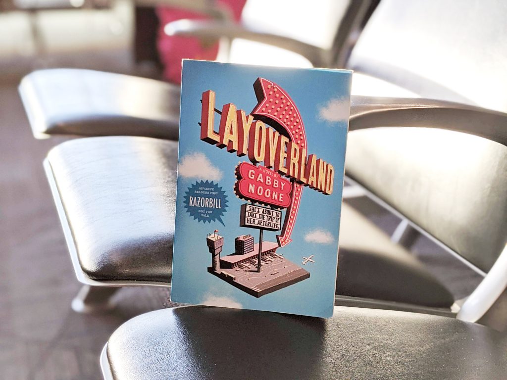 Layoverland by Gabby Noone sits on seats at an airport